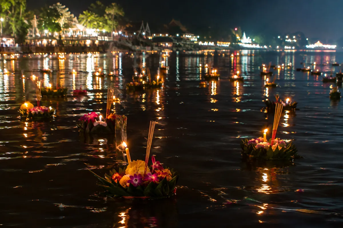 Flytande krathong av bananblad och blommor med tända ljus på en flod på kvällen
